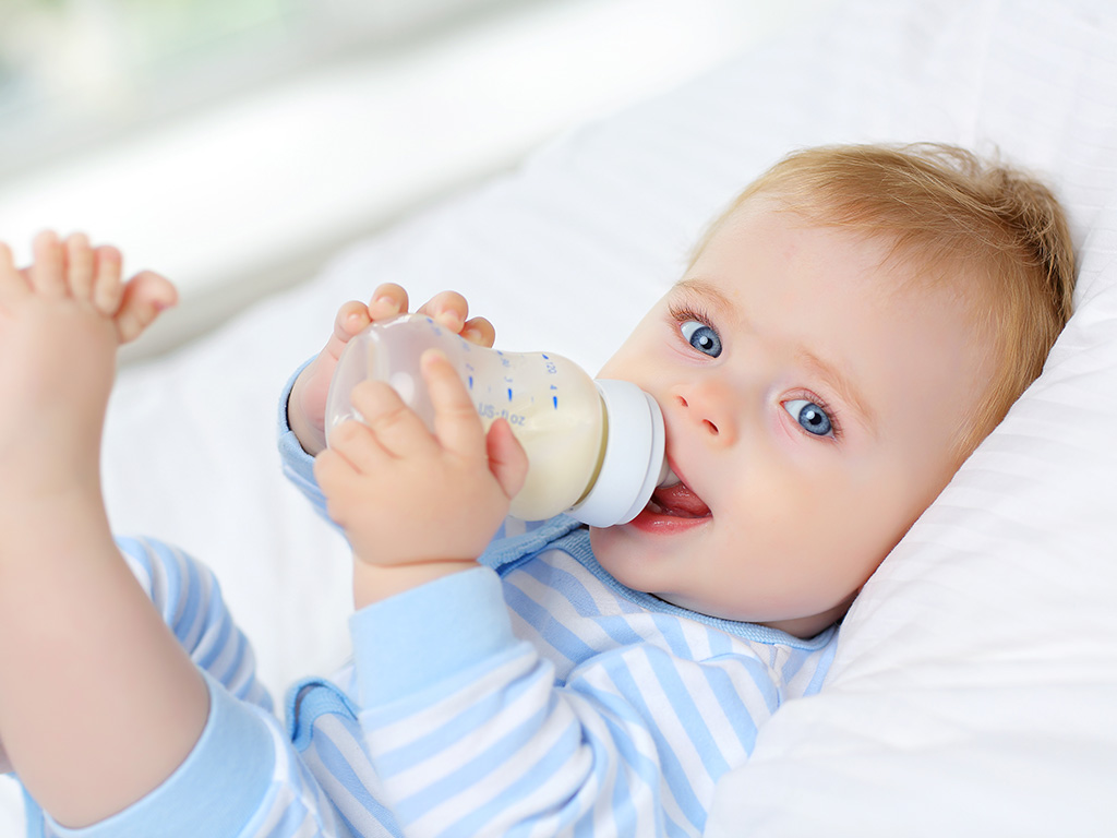 Charming blue-eyed baby 7 month old lies in bed and drinks milk from a bottle
